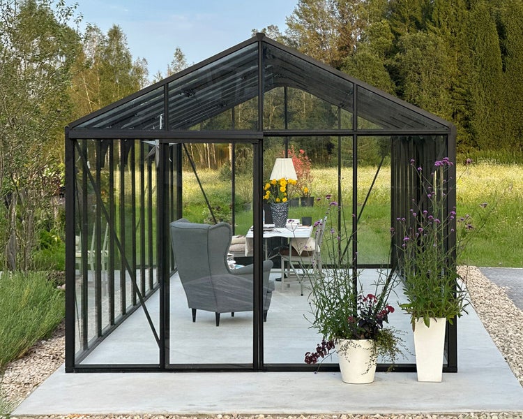 Gewächshaus mit schwarzem Metallrahmen als Wohnraum im Garten mit Sessel, Tisch und Pflanzen auf einer Betonplatte vor Waldkulisse.