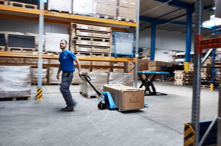 Ein Mitarbeiter zieht einen blauen Hubwagen mit einem großen Karton durch ein helles Lager mit hohen Regalsystemen.