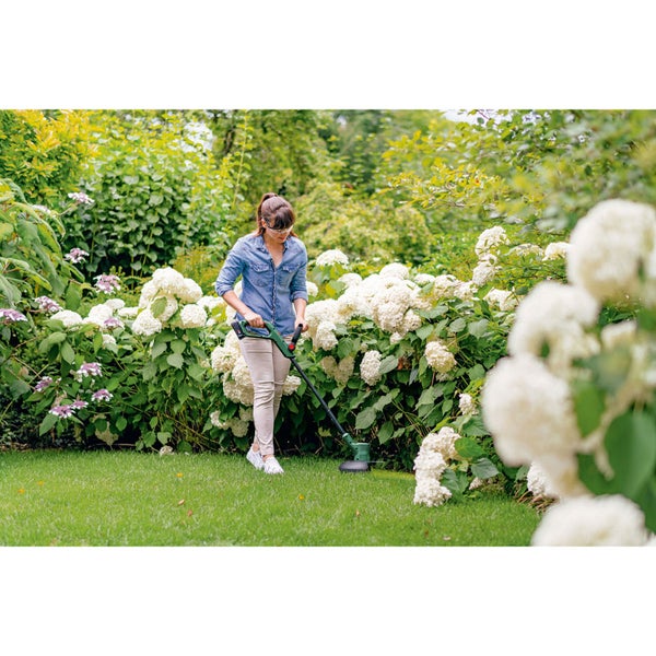 Eine Frau kürzt mit einem grünen Rasentrimmer die Rasenkante in einem hellen Garten mit blühenden weißen Hortensien.