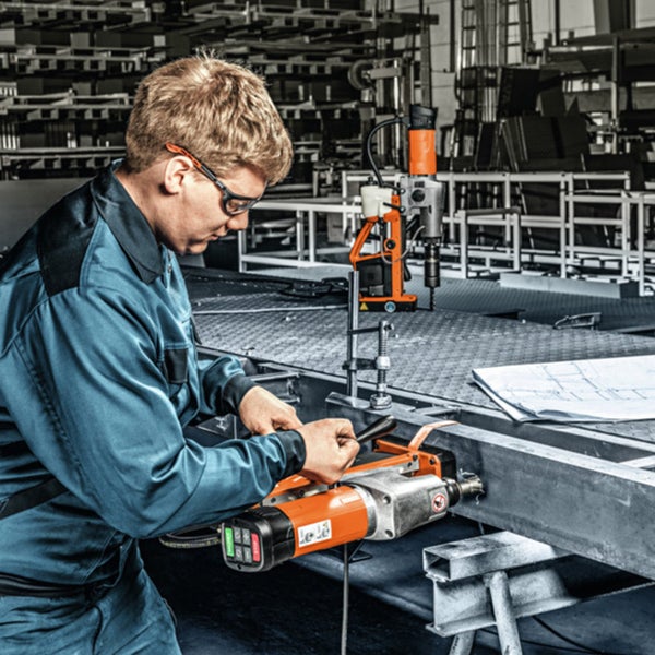 Ein Handwerker mit Schutzbrille bohrt mit einer Kernbohrmaschine in einen Metallträger in einer hellen Werkstatt.