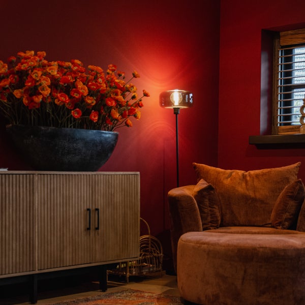 Wohnzimmer mit Kommode, Blumenarrangement, Stehlampe und Sessel vor einer roten Wand