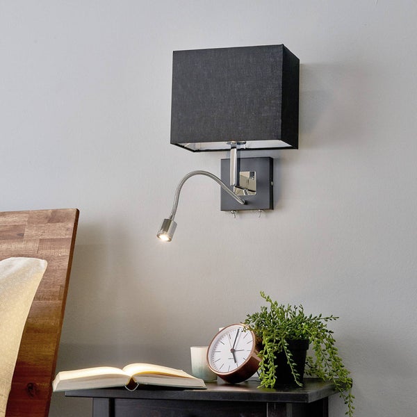 Wandleuchte mit schwarzem Stoffschirm und LED-Lesearm, montiert über einem Nachttisch mit Buch und Pflanze in einem hellen Schlafzimmer.