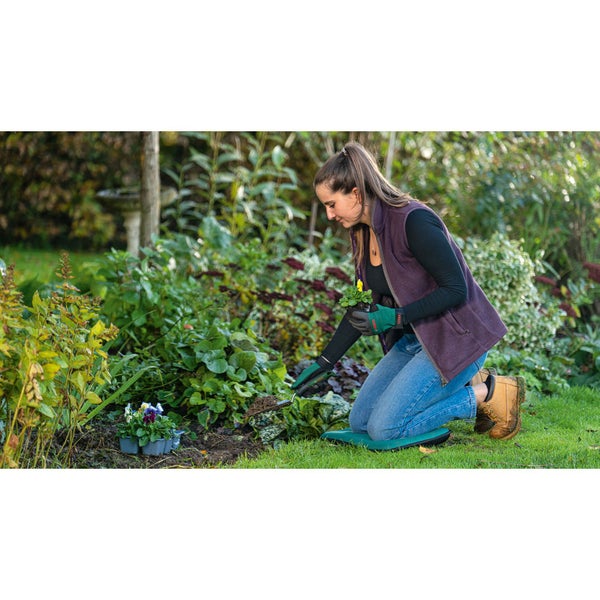 Frau kniet mit Gartenhandschuhen und Pflanzkissen im Garten und pflanzt Blumen.