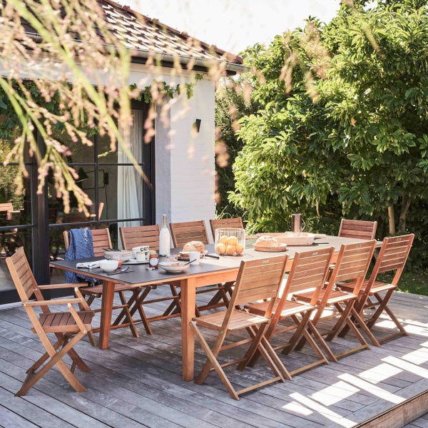 Gedeckter Gartentisch aus Holz mit acht passenden Klappstühlen auf einer Holzterrasse in einem hellen Garten neben einem Haus.