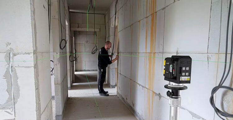 Handwerker richtet an einer Porenbetonwand einen Kreuzlinienlaser auf einem Stativ aus, der eine grüne Laserlinie im Rohbau projiziert.
