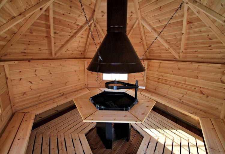 Innenansicht einer Grillkota aus Holz mit zentraler Grillstelle, Rauchabzugshaube und umlaufenden Holzbänken.