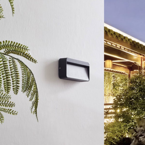 Schwarze, rechteckige LED-Außenwandleuchte im modernen Design an einer weißen Wand neben Farnblättern.