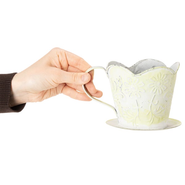 Hand hält einen dekorativen Pflanztopf aus Metall in Tassenform mit Henkel und floralem Relief.