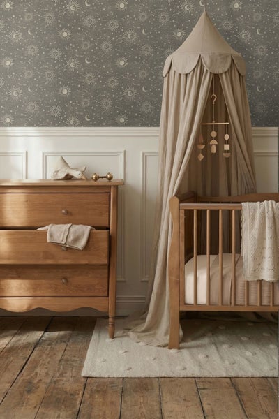 Kinderzimmer mit Vliestapete mit Sonnen, Mond und Sternen, Holzkommode, Gitterbett mit Baldachin und weißer Wandvertäfelung.