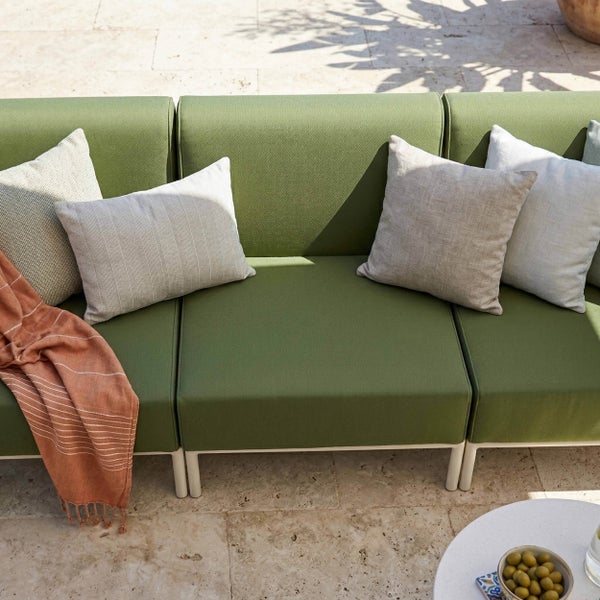 Grünes Gartensofa mit hellgrauen Zierkissen und einem gestreiften Plaid auf einer hellen Steinterrasse.