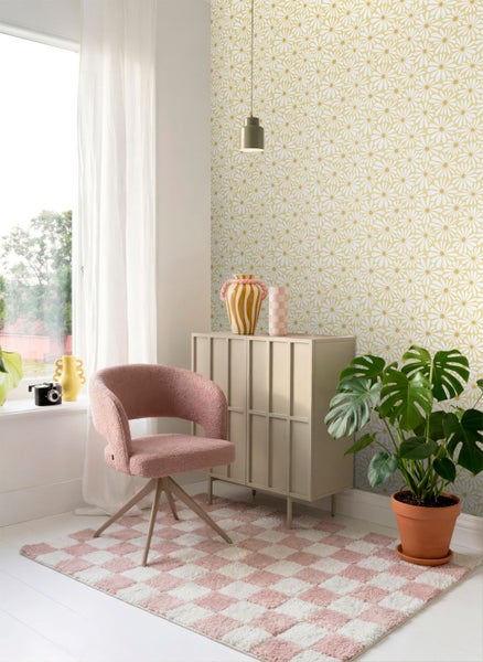 Hell gestaltetes Zimmer mit floraler Mustertapete, rosa Sessel, kariertem Teppich, beigem Schrank und einer Monstera-Pflanze.