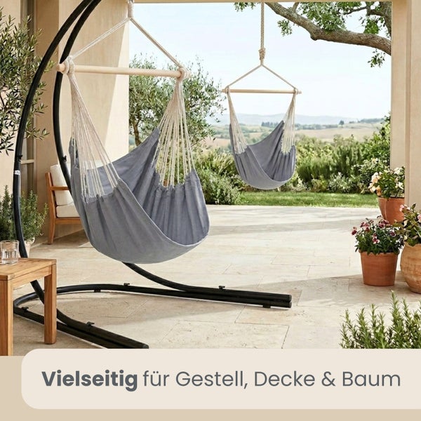 Zwei graue Hängesessel aus Stoff mit Spreizstab aus Holz auf einer Terrasse. Einer an einem schwarzen Metallgestell, der andere an einem Baum.