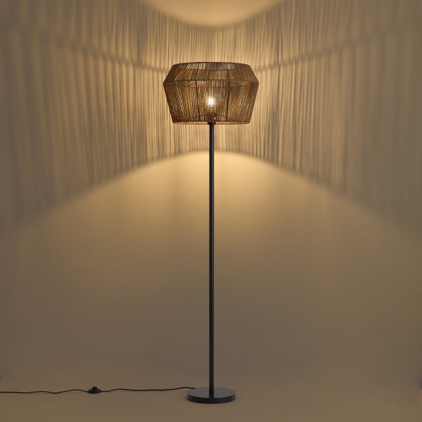 Stehlampe mit geflochtenem Schirm aus Seil auf schwarzem Metallgestell, wirft dekorative Schatten an die Wand.