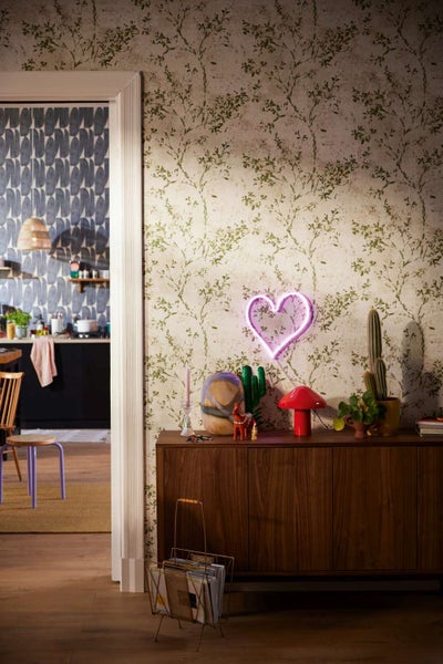 Wohnzimmer mit Vliestapete mit Pflanzenmotiv, Neonherz an der Wand, Sideboard aus Holz, Kakteen und Pilzlampe in einem modern eingerichteten Raum.
