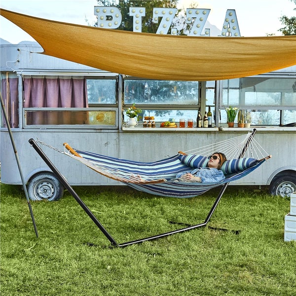 Gestreifte Hängematte mit Metallgestell und Kissen auf einer Wiese, im Hintergrund ein Wohnwagen unter einem Sonnensegel.