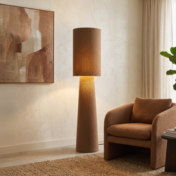 Moderne Stehlampe mit zylindrischem Schirm und konischem Fuß in texturierter Optik neben einem Sessel in einem wohnlich eingerichteten Zimmer.