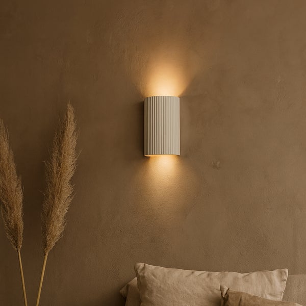 Moderne Wandleuchte mit geripptem Design und indirektem Lichtkegel nach oben und unten an einer Wand mit dekorativen Gräsern.