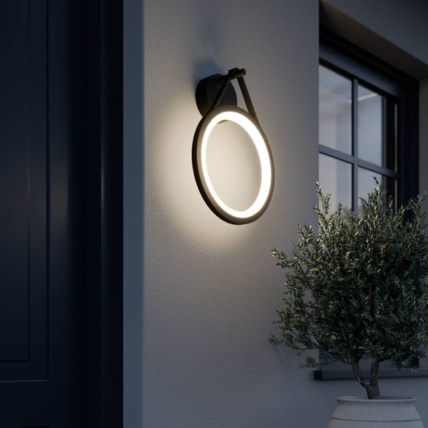 Moderne ringförmige LED-Außenwandleuchte in Schwarz an einer Hauswand neben einer Tür und einer Pflanze.