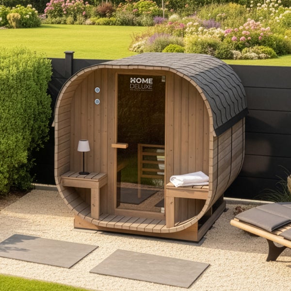 Fasssauna aus Holz mit Glastür und Home Deluxe Logo im Garten auf einem Kiesplatz mit zwei Sitzbänken vor dem Eingang.