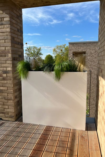 Großer rechteckiger weißer Pflanzkübel auf einem Holzbalkon mit Gräsern und Blumen vor gemauerten Wänden und blauem Himmel.