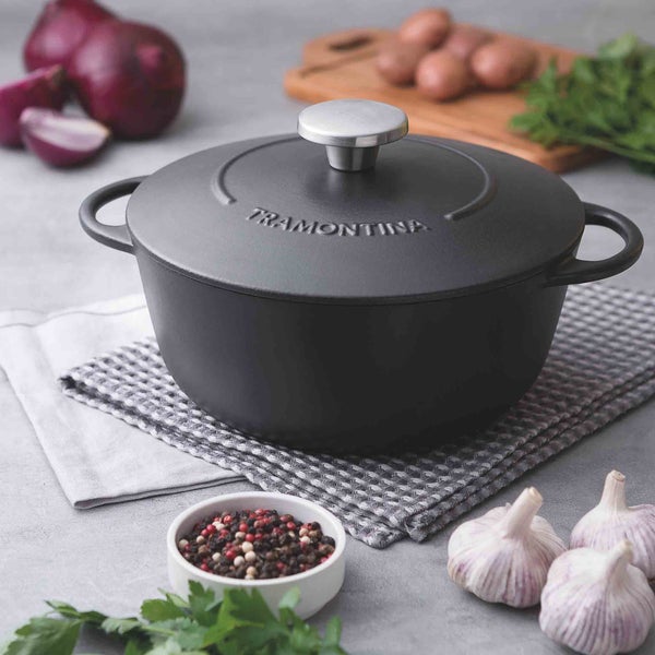 Schwarzer Tramontina Kochtopf aus Gusseisen mit Deckel auf einem Küchentuch mit Zwiebeln, Knoblauch, Kräutern und Gewürzen