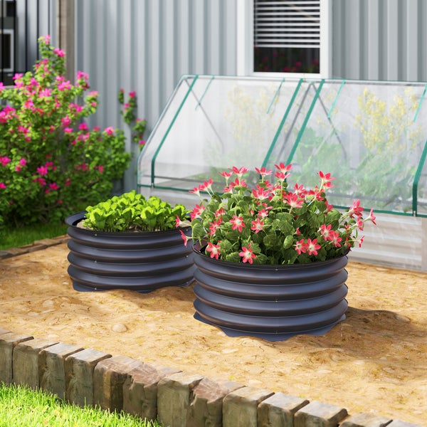 Zwei runde Hochbeete aus gewelltem Metall im Garten, bepflanzt mit rosa Blumen und Grünpflanzen, im Hintergrund ein Kleingewächshaus.