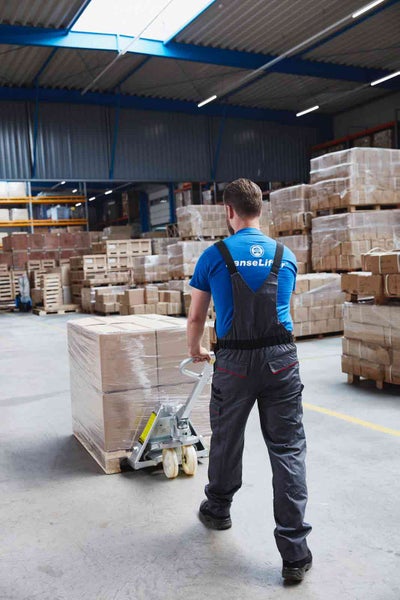 Ein Mitarbeiter in grauer Latzhose und blauem T-Shirt mit HanseLif Logo zieht einen beladenen Hubwagen durch ein helles, organisiertes Lager.