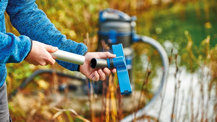 Eine Person montiert eine blaue Saugdüse an ein silbernes Saugrohr eines Schlammsaugers an einem hellen Gartenteich.