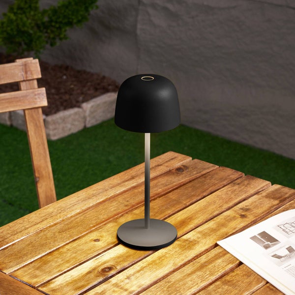 Moderne kabellose LED-Tischleuchte auf einem hölzernen Gartentisch im Außenbereich mit schwarzem Schirm und schlankem Standfuß.