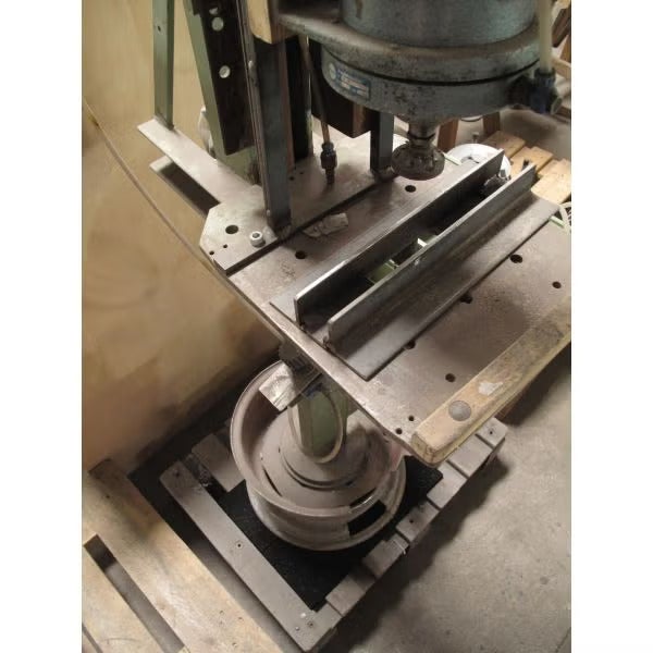 Eine Ständerbohrmaschine oder Presse in einer Werkstatt, montiert auf einem stabilen Tisch mit Metallschienen, stehend auf einer Holzpalette.
