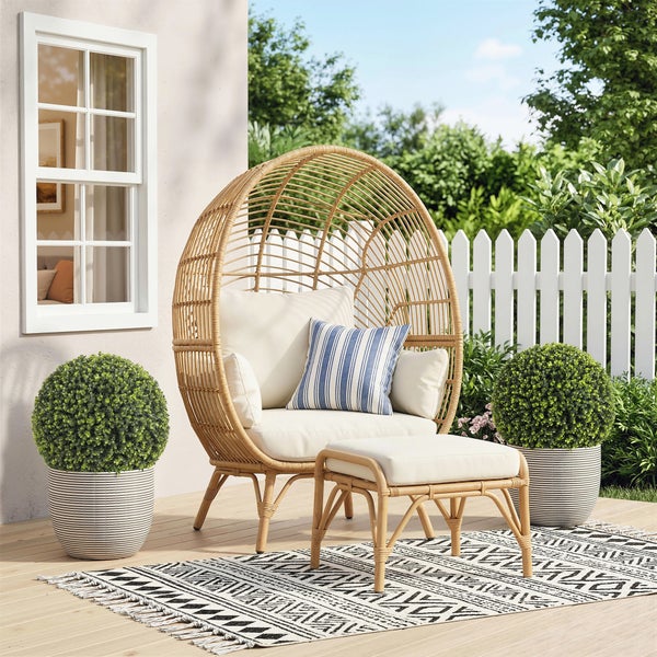 Garten-Loungesessel in Eiform aus Korbgeflecht mit passendem Fußhocker, hellen Sitzpolstern und gemustertem Outdoorteppich auf einer Terrasse.
