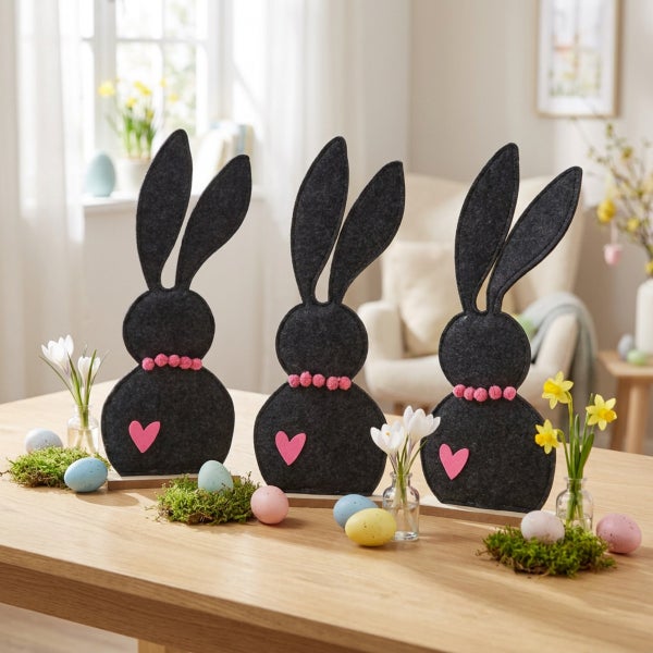 Drei Osterhasen Figuren aus Filz als Tischdekoration mit Herz Applikation und Frühlingsblumen auf einem Holztisch.
