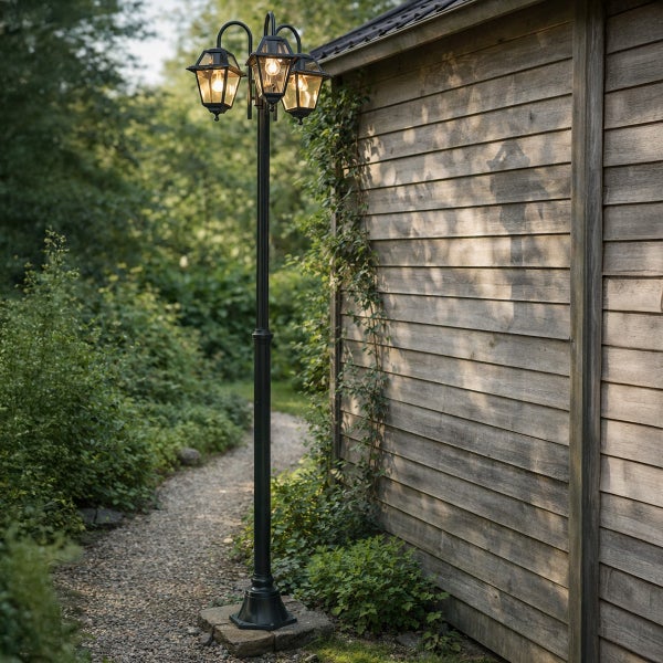 Dreiflammige Gartenmastleuchte in klassischem Stil neben einer Holzfassade auf einem Kiesweg.