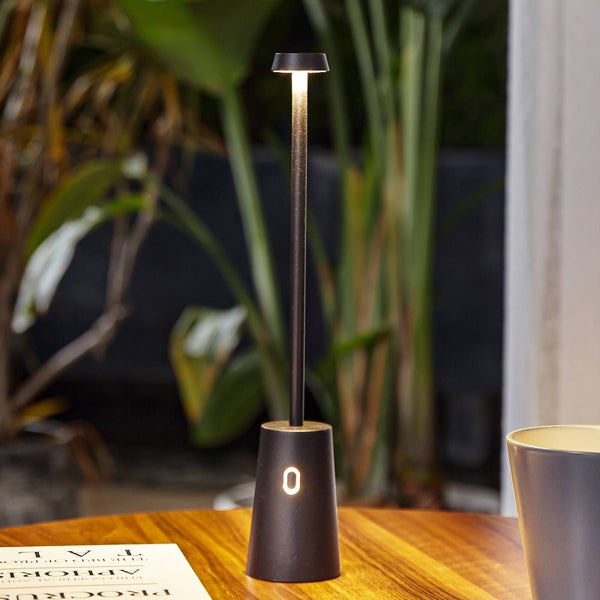 Tragbare LED-Tischleuchte mit schlankem Stab und konischem Fuß auf einem Holztisch vor Pflanzen im Hintergrund.
