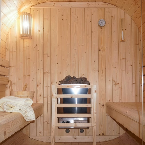 Innenansicht einer Fasssauna aus Holz mit Saunaofen, Saunabänken, Lampe, Thermometer und Hygrometer für ein entspannendes Wellnesserlebnis.