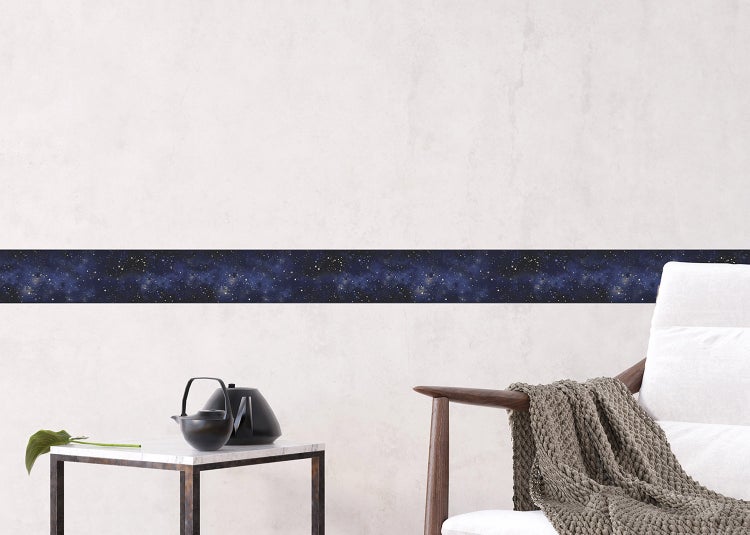 Selbstklebende Tapetenbordüre mit dunkelblauem Sternenhimmel-Motiv an einer hellen Wand in einem hellen Wohnzimmer mit Sessel und Beistelltisch.