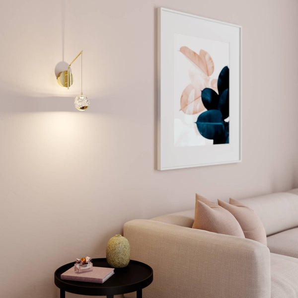Moderne Wandleuchte mit goldfarbenem Arm und Glaskugel in einem hellen Wohnzimmer neben einem Sofa und einem eingerahmten Bild.