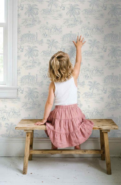 Kind sitzt auf einer Holzbank vor einer Vliestapete mit Safari-Motiv im Toile-de-Jouy-Stil in einem hellen Kinderzimmer.