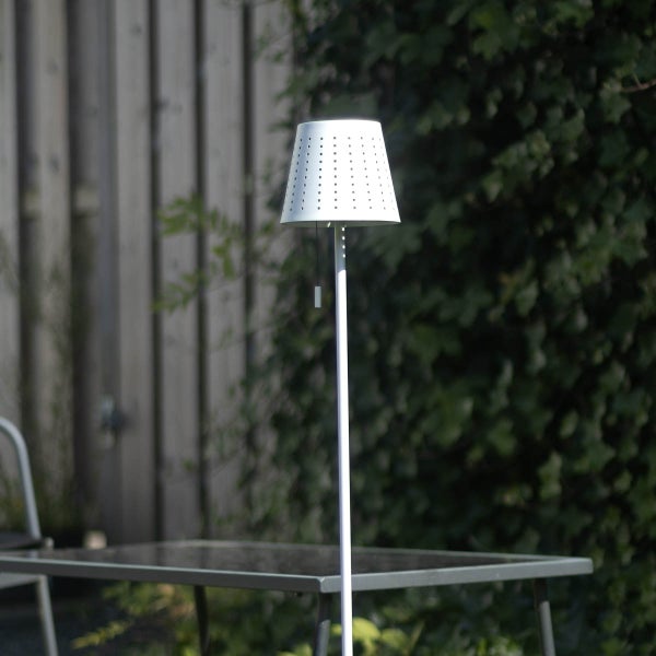 Solar-Stehleuchte für den Außenbereich mit weißem, perforiertem Schirm, platziert in einem Garten neben einem Tisch.