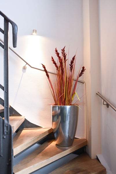 Moderne Bodenvase aus Metall mit hohem Ziergras in Rotbraun auf einer Holztreppenstufe mit Metallhandlauf.