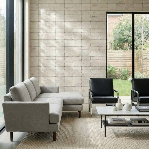 Modernes Wohnzimmer mit heller Ziegelwand, grauem Ecksofa, zwei Ledersesseln, Marmor-Couchtisch und großem Fenster mit Blick in den Garten.