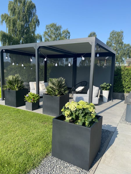 Moderne Terrasse mit schwarzer Lamellen-Pergola, Lounge-Möbeln, großen schwarzen Pflanzkübeln und Lichterkette auf grauen Bodenplatten neben Rasen.