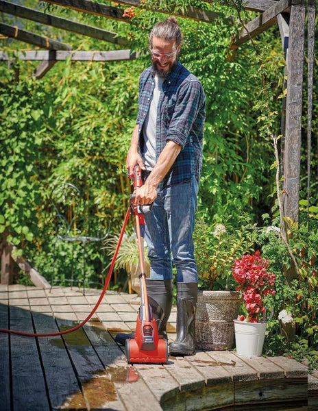 Ein Mann reinigt mit einer roten elektrischen Oberflächenbürste eine Holzterrasse in einem hellen Garten mit Pflanzen und Blumen.