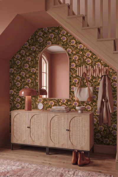 Ein Eingangsbereich unter einer Treppe mit floraler Vliestapete, einem Sideboard aus Holz mit Wiener Geflecht und einem gewölbten Wandspiegel.
