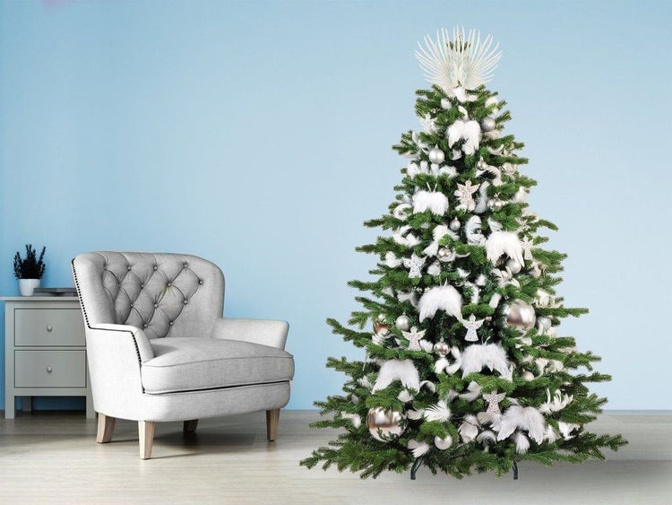 Ein geschmückter künstlicher Weihnachtsbaum mit silbernem Schmuck, weißen Federn und einer Baumspitze, daneben ein Polstersessel und eine Kommode.