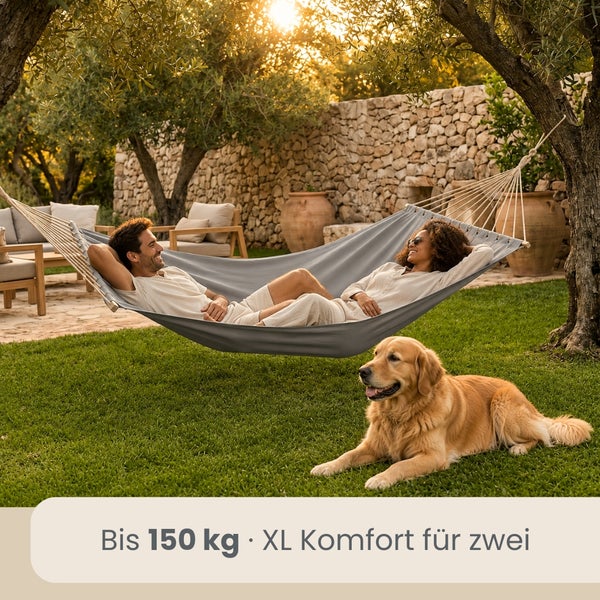 Große Hängematte im hellen Garten, Belastbarkeit bis 150 Kilogramm, XL Komfort für zwei Personen, Szene mit Steinmauer, Bäumen und einem Hund.