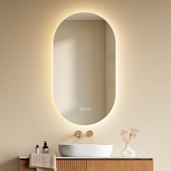 Ovaler Badspiegel mit LED-Beleuchtung und Touch-Bedienung über einem weißen Waschbecken mit goldener Armatur und Holzunterschrank.