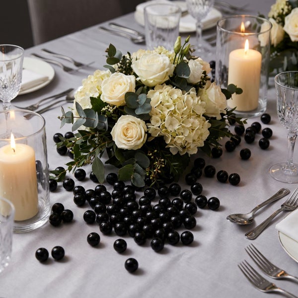 Festlich gedeckter Tisch mit Blumengesteck aus Rosen und Hortensien, Stumpenkerzen in Glaszylindern und dekorativen dunklen Kugeln als Tischschmuck.
