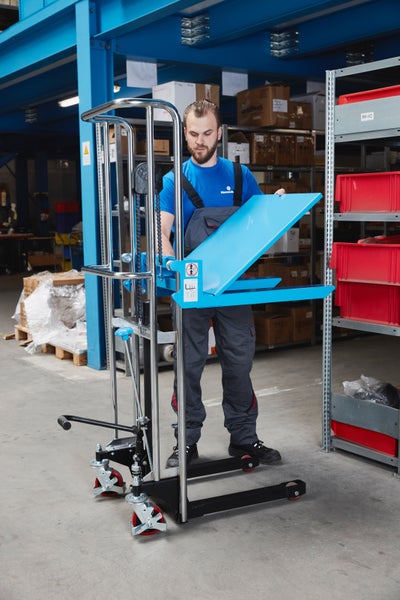 Ein Lagerarbeiter bedient einen manuellen Hochhubwagen mit einer blauen kippbaren Plattform in einem Lager.