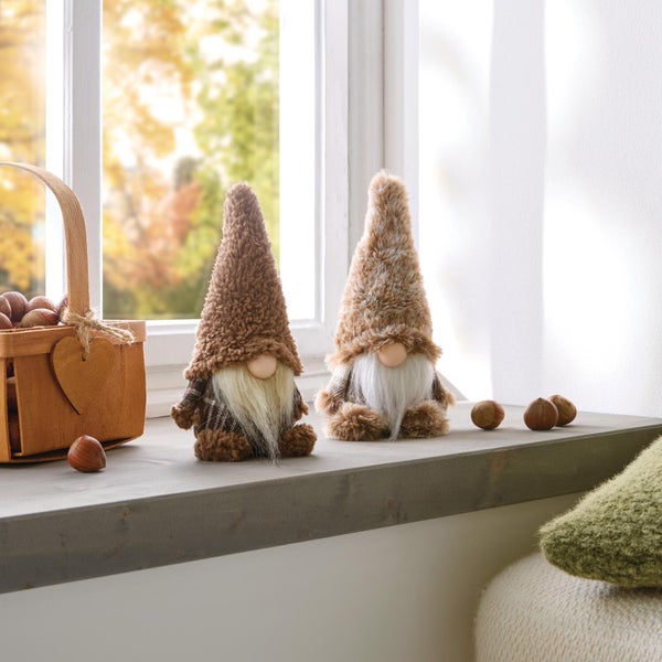 Zwei dekorative Wichtelfiguren mit flauschigen Mützen und Bärten auf einem Fensterbrett neben einem Holzkorb mit Haselnüssen.